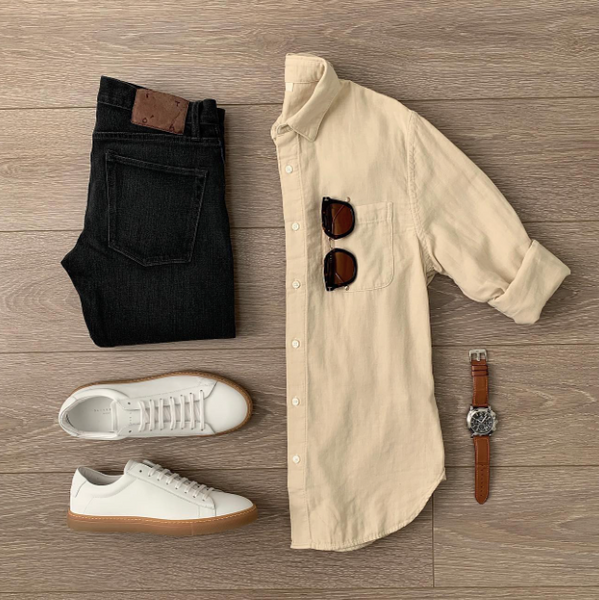 On a wooden background. A beige shirt, black jeans, and white sneakers, along with a brown watch and glasses