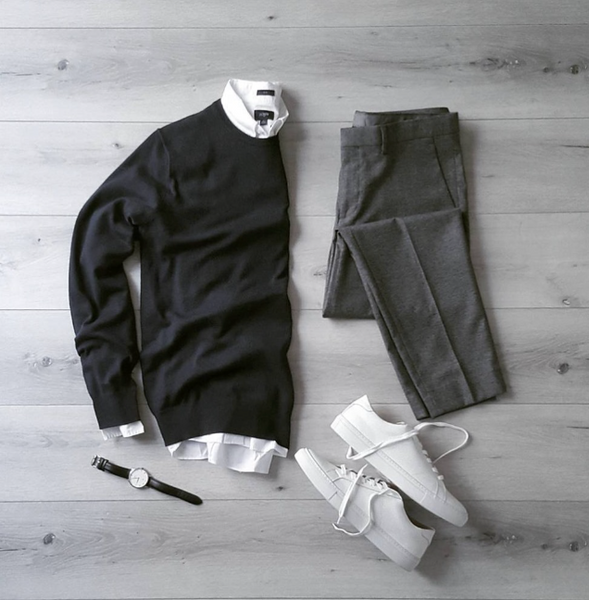 On a wooden background; A black sweater, over a white shirt. White sneakers, a black belt, and gray chinos 