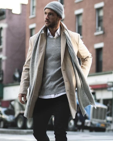Man in the city wearing black pants, a white button-down, gray sweater, beige duster, gray beanie & gray scarf