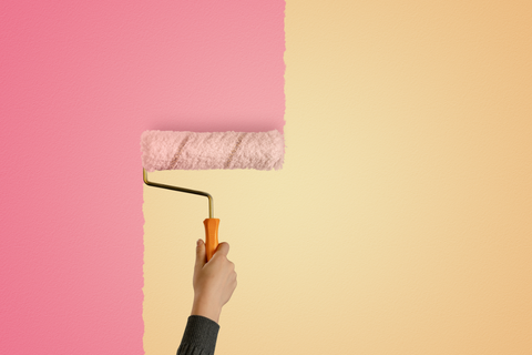 Hand holding a rolling paint brush and painting pink paint over a burnt yellow colored wall.