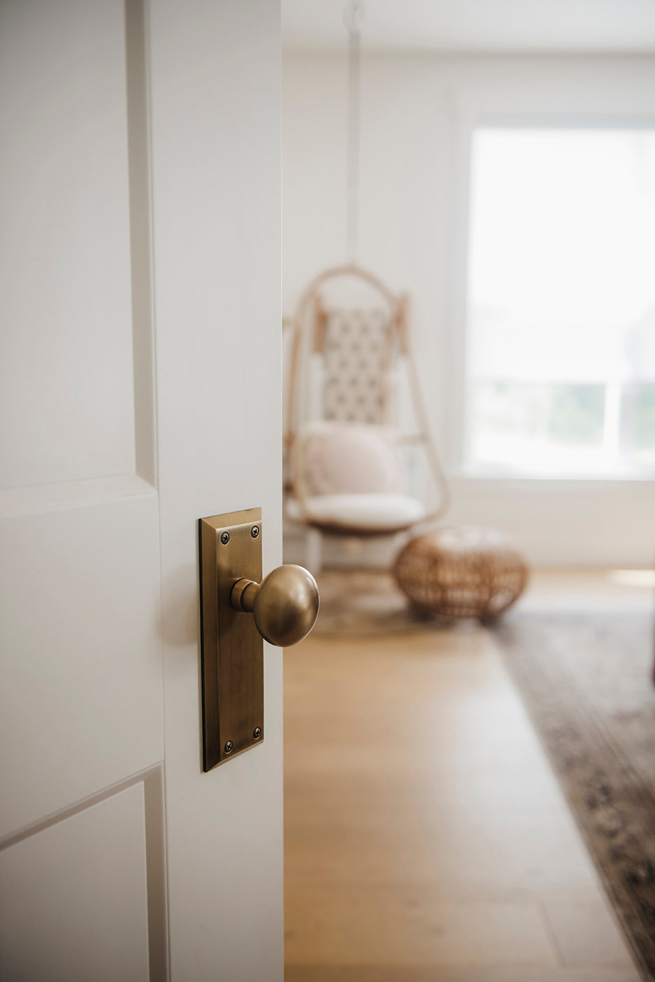 White door opening into a living space with a hanging chair in the background. The door has a Fifth Avenue Long Plate with Fifth Avenue Knob in Vintage Brass door knob.