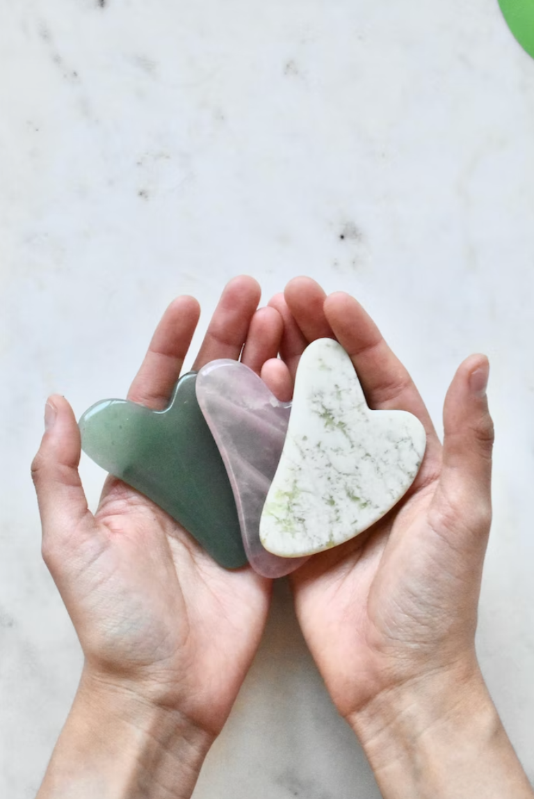 a person's hands holding three gua sha tools in jade, rose quartz and marble