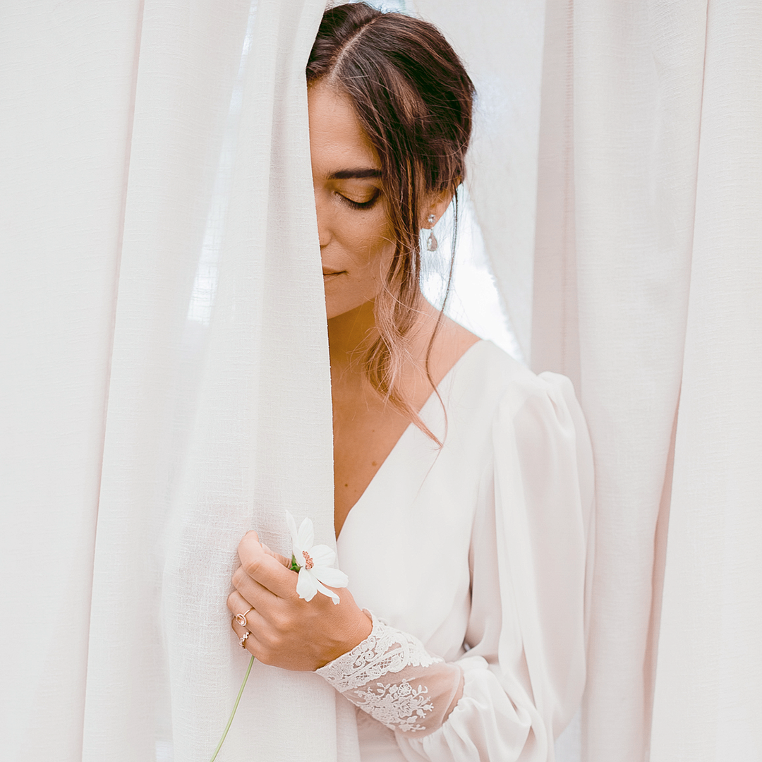 A bride in a white sleeved wedding dress holding a white flower and half hidden behind a sheer white curtain