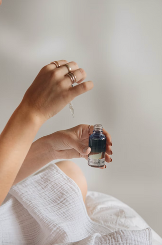 Image by Mathilde Langevin via Unsplash, pictured is a woman in a white linen dress holding a serum vial for oils