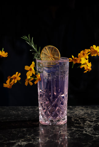 A purple gin and tonic is garnished with yellow flowers and a slice of dried orange. 