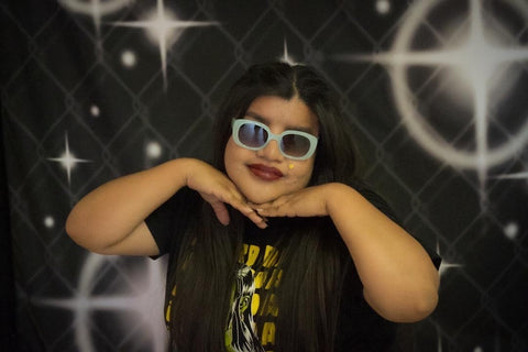 Owner, Edith Perez, posing with hands under chin, in front of a classic 90s backdrop of 4 point stars scattered through. She is wearing cool blue sunglasses and a dark red lip, with long black hair and wearing the Mordida shirt