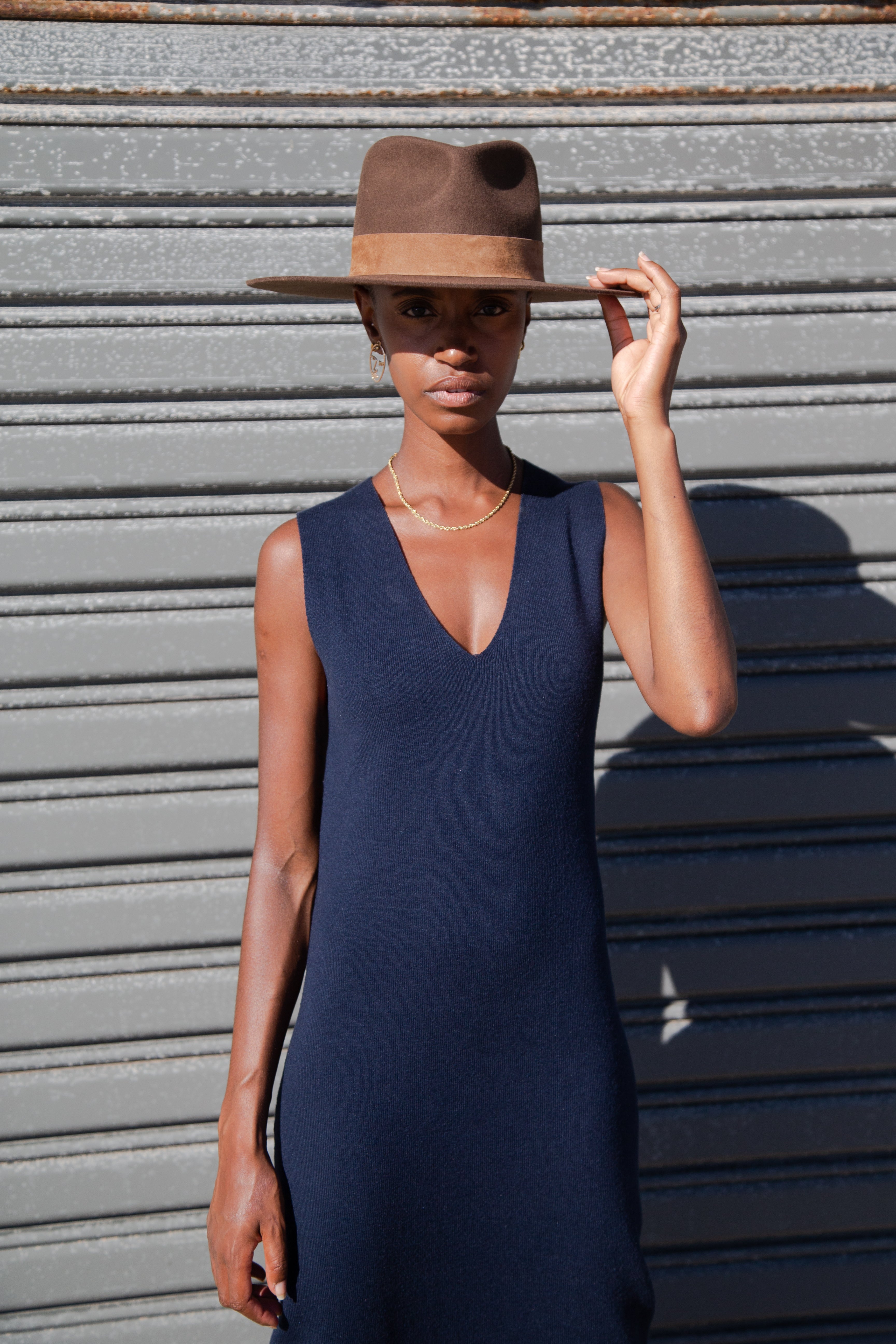 Sinead is looking straight at the camera holding the tip of her Brown Lack of color hat.  She is wearing the Havana knit dress in midnight Navy with a gold chain around her neck,