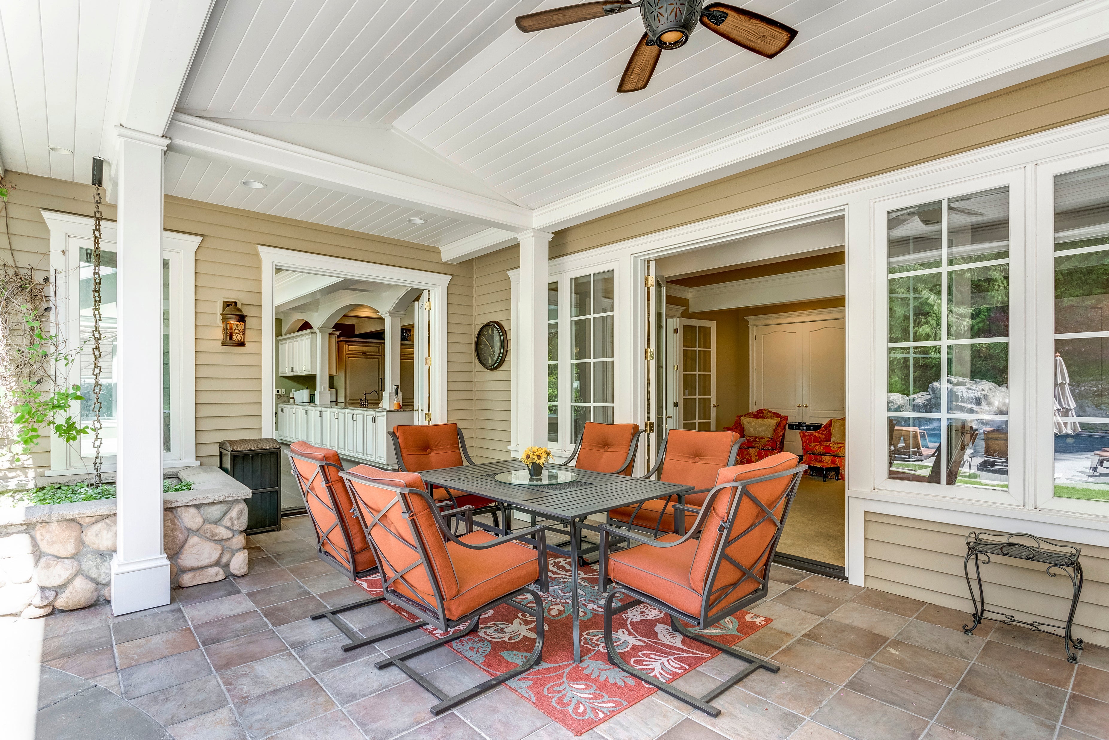 Back porch of a white house with a dining table set that is dark metal and orange pillows.