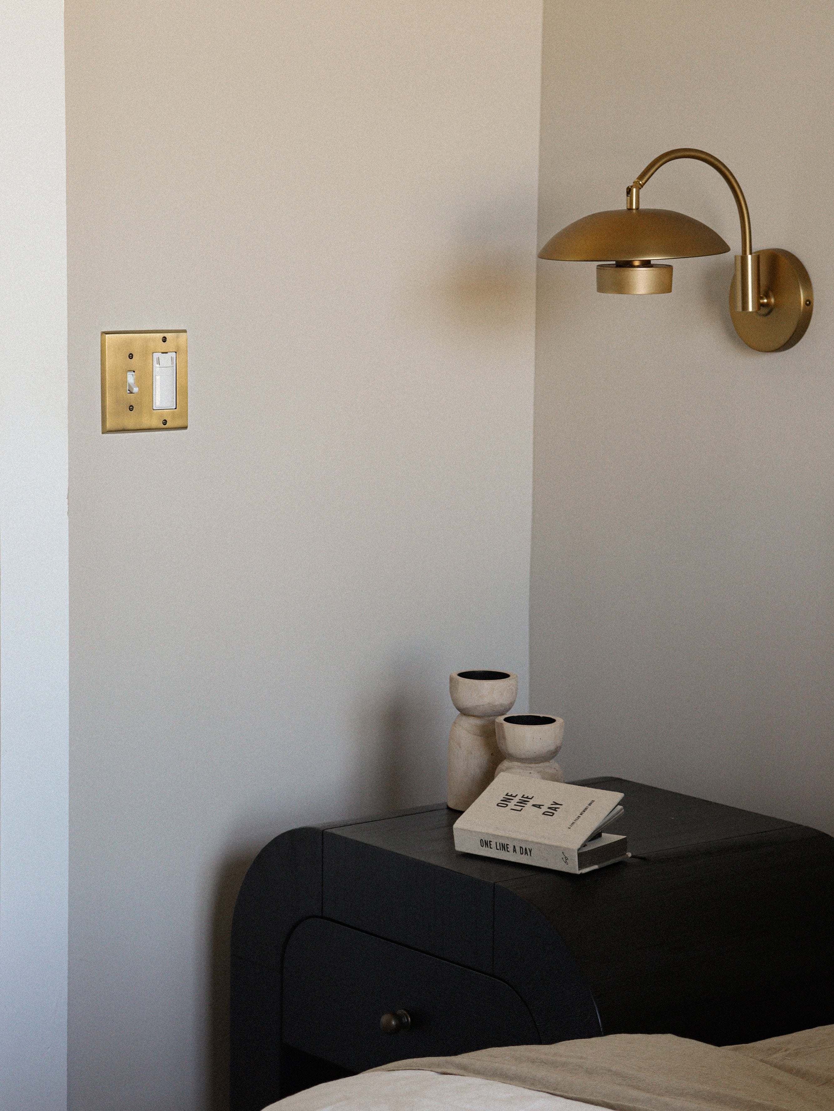 Bedside table in a corner with a lamp over it and a New York Switch Plate with Toggle and Rocker in Polished Brass on the other wall.