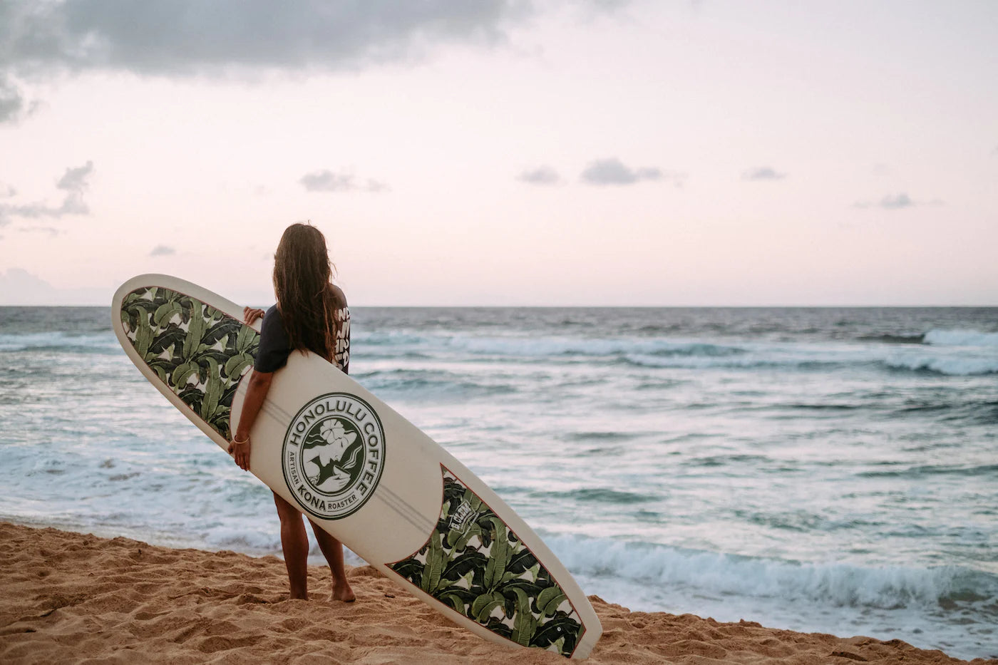 Woman holding custom Honolulu Coffee surfboard at the beach