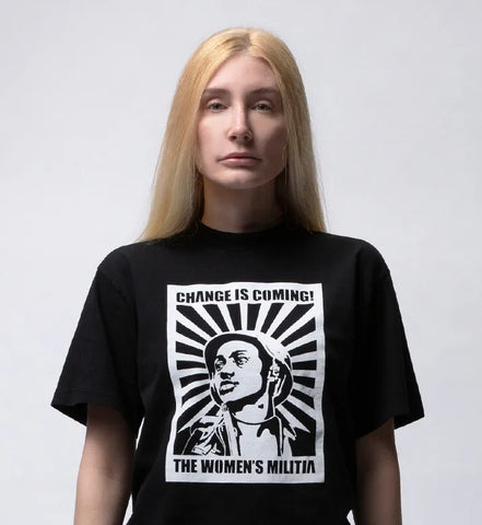 Portrait photo of Artist Leigh Brooklyn, looking forward wearing a black t-shirt with a graphic of a military woman on it. The shirt reads, Change is Coming, the Women's Militia