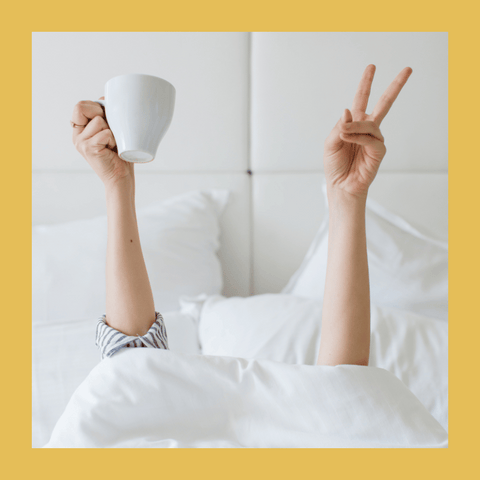 Parent in bed with both hands raised. One hand holding coffee cup. One hand with the peace sign.