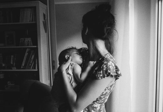 Mother holding sleeping baby in a dark room looking away from camera.