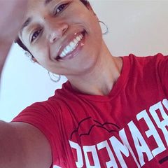 square photo of TaLisa in selfie style with a plain white/grey background. the image is tilted so that her head and body are slanted coming diagonally from the top left corner. she is wearing a bright red t-shirt with big white letters that are in mirror view and cropped (but says “DREAMERS”). she is smiling brightly.