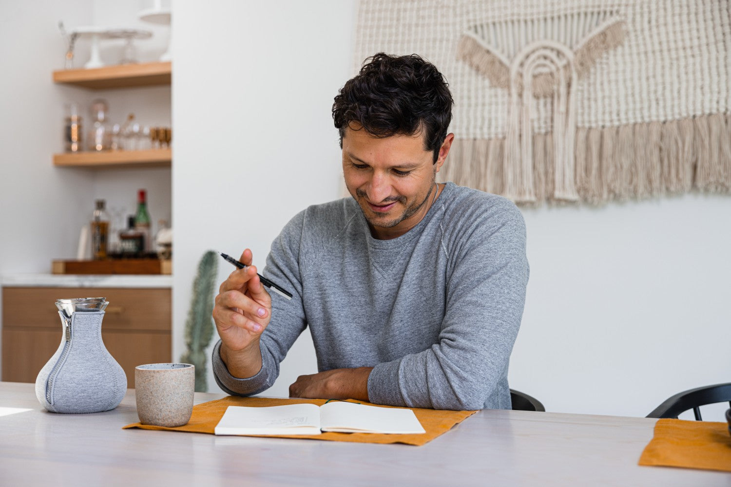 Man sitting at a table with a journal and coffee with macrame hanging behind him