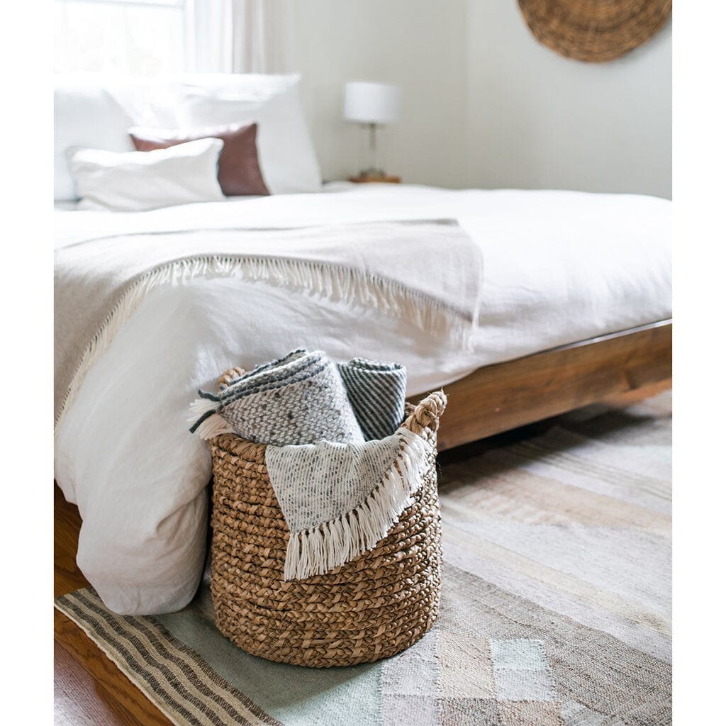 A woven basket at the base of a bed with cozy throw blankets