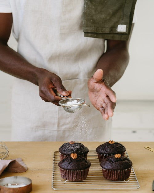 Person sprinkling sugar on cupcakes with tea towel on their shoulder