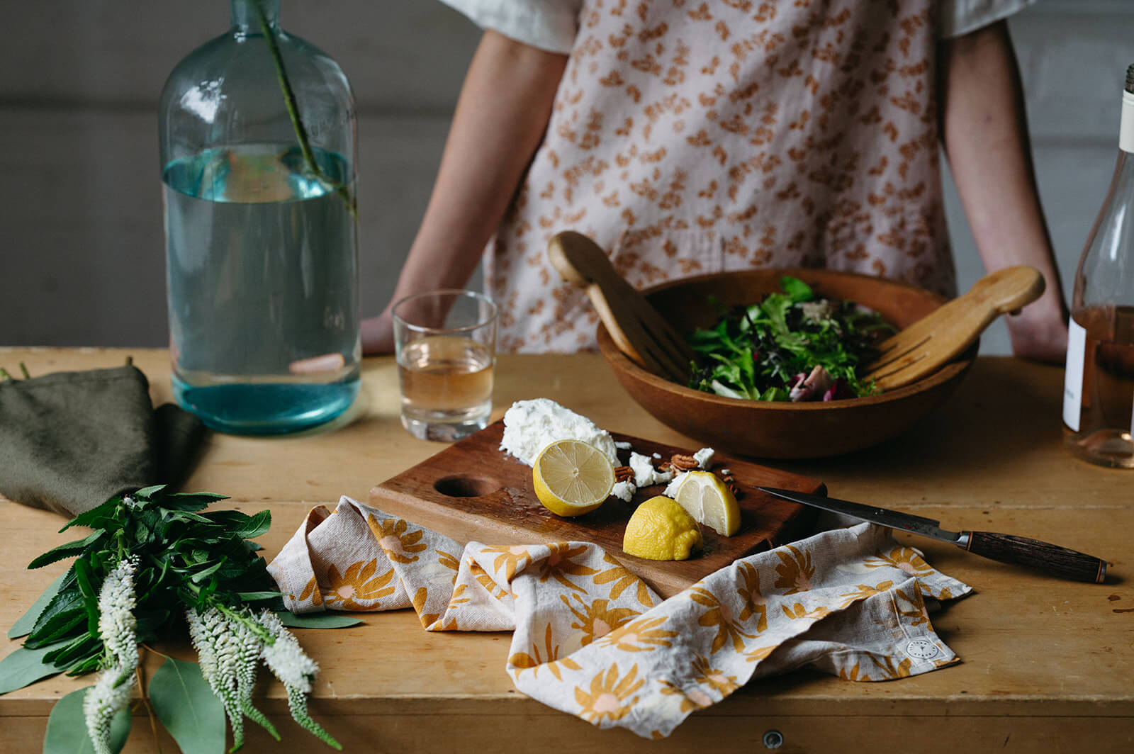 Wooden table with bowl of salad, lemons and cheese on a cutting board and a dish towel