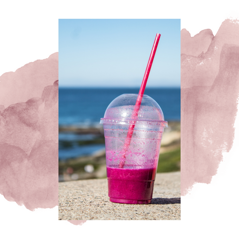 Plastic straw and plastic cup half filled with pink liquid, at the beach on a concrete side. 