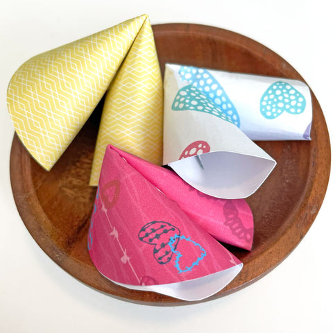 3 colorful patterned origami folded paper fortune cookies sitting on a small wooden plate on a white background. Learn to fold your own paper fortune cookies! Instructions and free printable design template by Sunny Day Designs