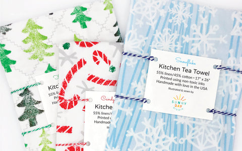 3 colorful, holiday printed fun tea towels by Sunny Day Designs are packaged on a white background. A green tree printed woodland dish towel named "Friendly Forest", a red candy cane printed kitchen towel named "Candy Cane Lane" & a light blue snow printed tea towel named "Snowflake". All 3 handmade tea towels are made in the USA from linen/cotton fabrics.