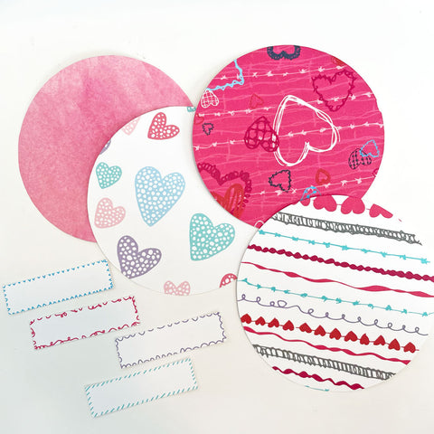 4 Colorful Round Circles with multicolored/pink valentine's day themed patterns and 4 rectangular paper strips sit on a white background. These supplies are used to create paper fortune cookies. Printable template and instructions by Sunny Day Designs.