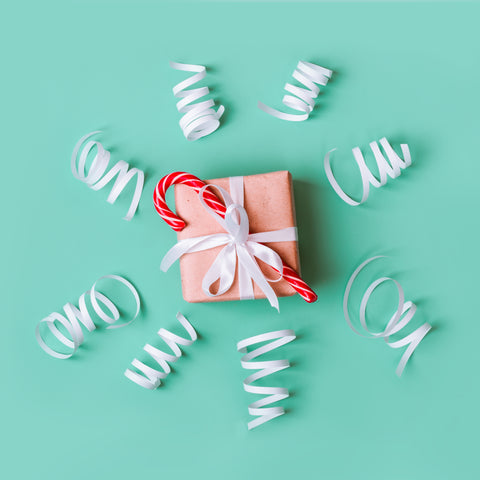 Blog post image for holiday shopping guide for conscious consumers by Sunny Day Designs. A square Christmas present box decorated with kraft paper, tied with white ribbon and topped with a candy cane. Curly white ribbon curls suround the holiday present box, which is placed on a solid light aqua background.