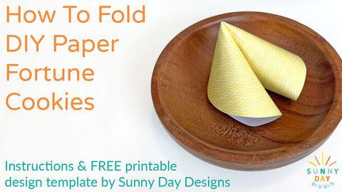 Yellow patterned paper fortune cookie in a wooden bowl on white table. Orange text reads "How To Fold DIY Paper Fortune Cookies". Turquoise text readsy Instructions & FREE design template by Sunny Day Designs". Click image for video instructions on how to fold paper fortune cookies!