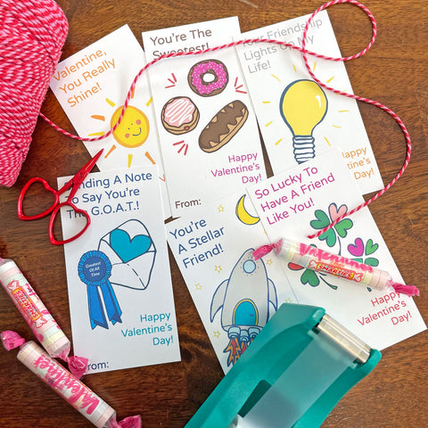 6 printable mini valentine cards (designed by Sunny Day Designs) have been cut out and are resting on a wood table alongside a spool of red/pink striped baker's twine, 3 wrapped candies, and a turquoise clear tape dispenser