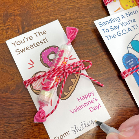 Hand with pen signing the name "Shelley" onto a free printable DIY valentine card with candy attached, artwork designed by Sunny Day Designs