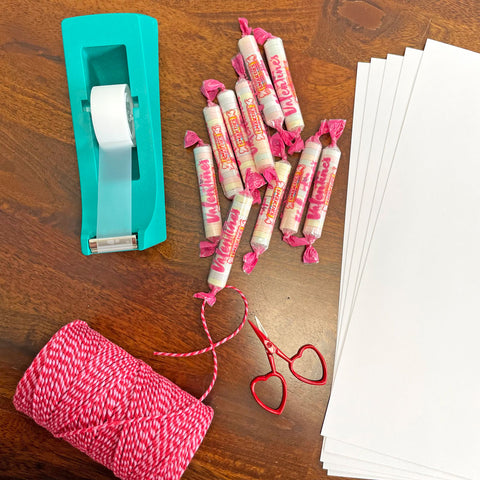 Items you'll need to create the printable valentines day cards by Sunny Day Designs are shown on a brown wood table: a turquoise tape dispenser, pink and red baker's twine, individually wrapped Smarties candies, sheets of white card stock paper