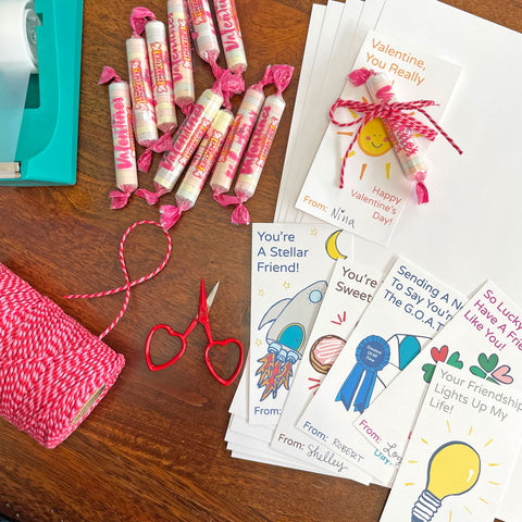 6 playful and colorful Free DIY printable valentines mini cards by Sunny Day Designs are shown on a wooden tabletop next to sheets of white card stock paper, a spool of red/pink striped baker's twine string, a turquoise clear tape dispenser, and a handful or wrapped candies