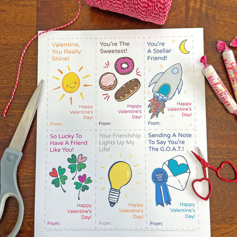 6 free playful printable valentines cards by Sunny Day Designs, uncut - shown on a brown wood table with a scissors, pink/red baker's twine spool, and wrapped candies