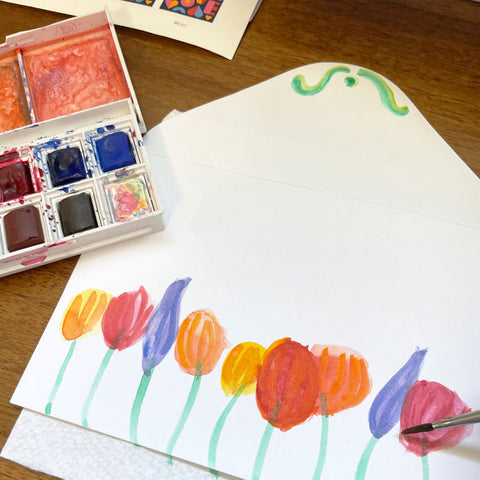 Colorful Watercolor Flowers Painted onto White Envelope Next to Watercolor Paint Set and on Wood Table. By Sunny Day Designs - Snail Mail Fun Blog Post