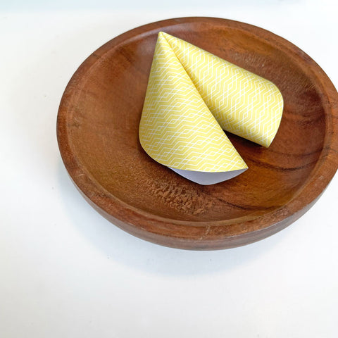 1 paper fortune cookie made out of patterned yellow paper rests inside a small wooden plate on top of a white table. Image by Sunny Day Designs