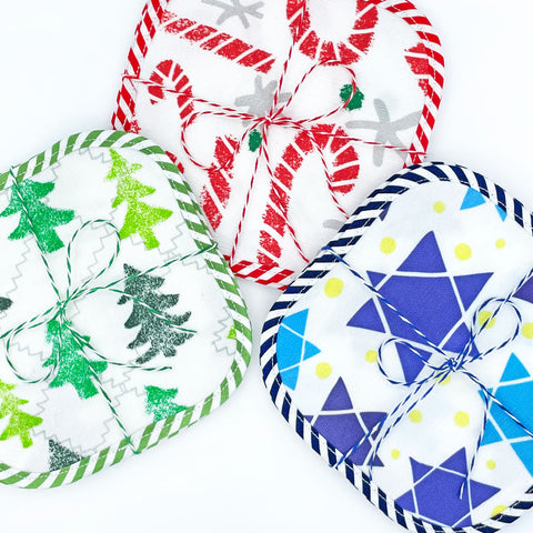 This images shows 3 sets of eco-friendly, square, washable fabric cocktail napkins in 3 holiday prints: Candy Cane Lane, Friendly Forest & Star of David. All cocktail napkins are made in the USA from a linen/cotton fabric. Prints are designed Sunny Day Designs.