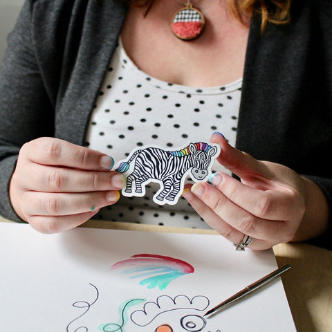 close up image of Shelley Schmidt holding a colorful zippy zebra vinyl sticker by Sunny Day Designs in front of an open sketchbook with drawings