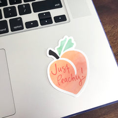 An orange, green, and coral "Just Peachy" vinyl sticker, designed by Sunny Day Designs, adhered to the right of a gray laptop track pad.