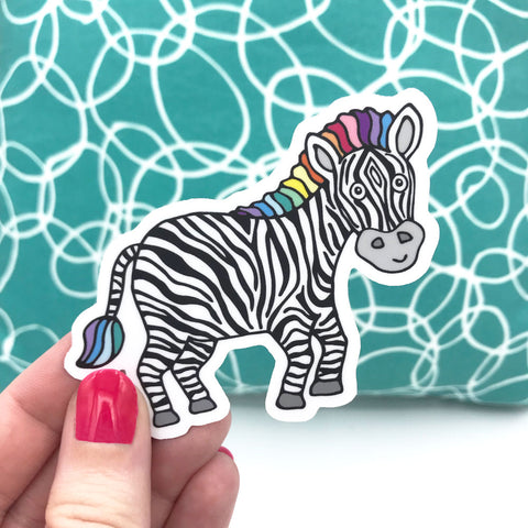 A colorful and cute zebra sticker is held in a hand in front of a turquoise and white printed pillow. This zebra sticker helps raise money for charity and supports the rare disease community.