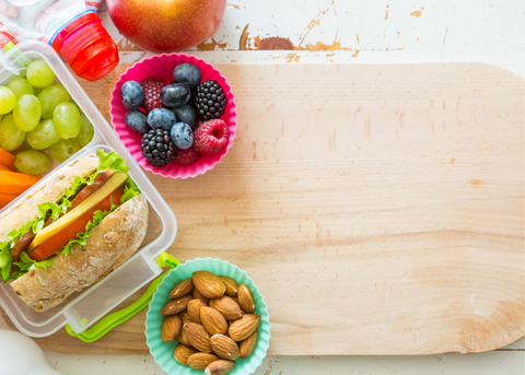 Packed lunch being prepared on a wood cutting board featuring a water bottle, sandwich, fruits and almonds.