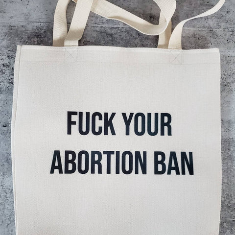 Natural colored faux burlap polyester linen tote bag with "FUCK YOUR ABORTION BAN" printed in black capital block lettering