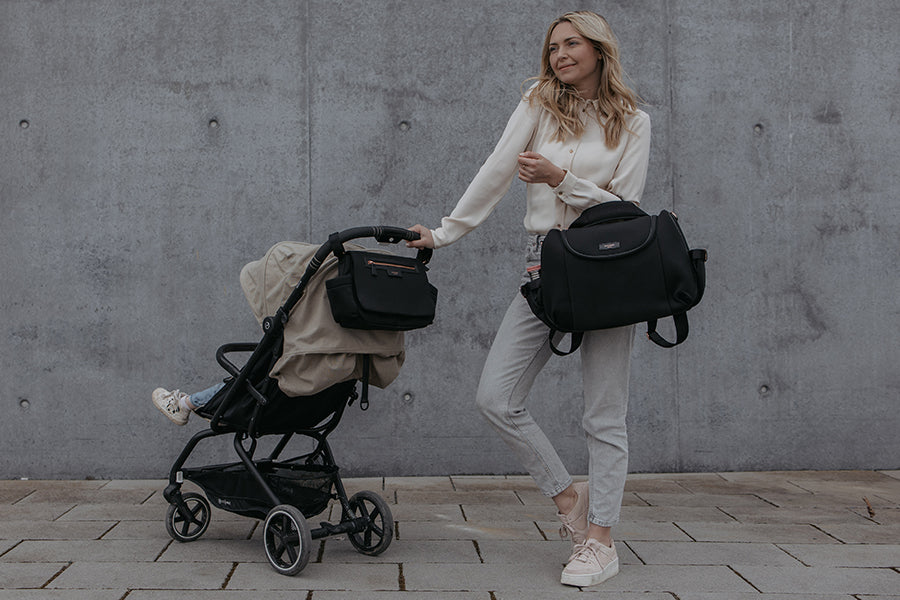Mum out with her stroller organiser luxe black on the stroller with the baby. She is also carrying her Poppy Luxe black changing bag as a convertible bag over her shoulder.