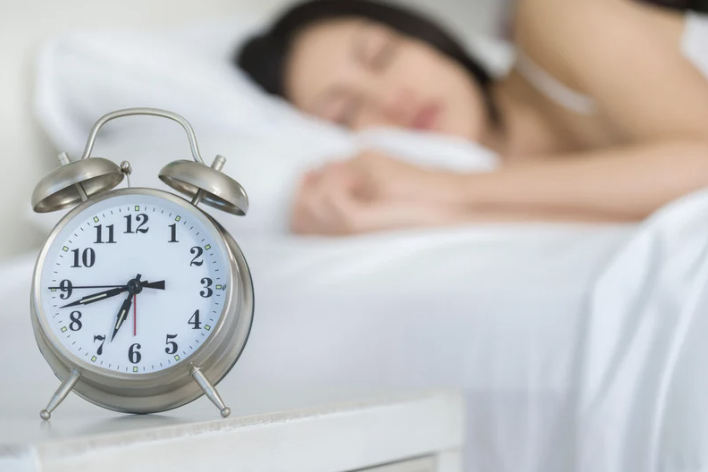 A clock in the foreground and a woman sleeping in bed enjoying the benefits of taking magnesium at night.