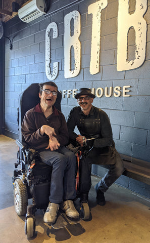 DrDazz, in his electric wheelchair, sitting next Vincent, who is wearing a leather rimmed hat. They next to a black painted brick inside wall with the letters CBTB painted on it. 