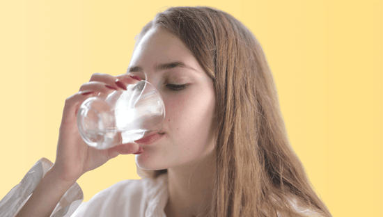 Young brunette woman with long hair in profile drinking a glass of water on yellow background