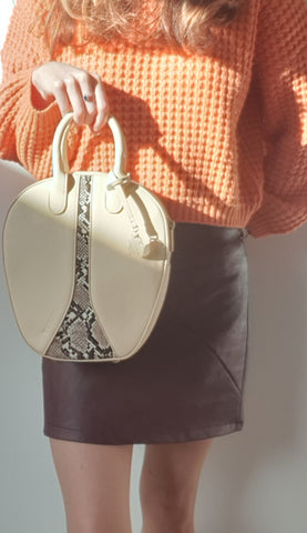 A women wears a orange knit jumper and burgundy vegan leather skirt.  She holds a cream and snake-skin patterned vegan recycled PU handbag in front of her.