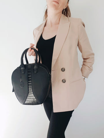 Women wearing a camel blazer and black trousers holds an oval black vegan leather shoulder bag.