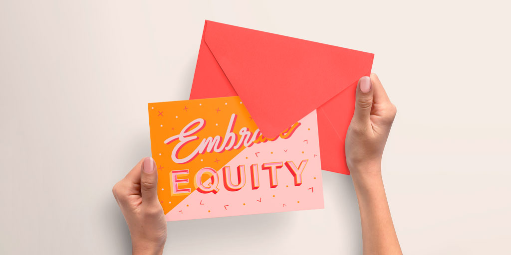 A women is holding an orange and baby pink card that reads Embrace Equity, the theme of International Women's Day in 2023.