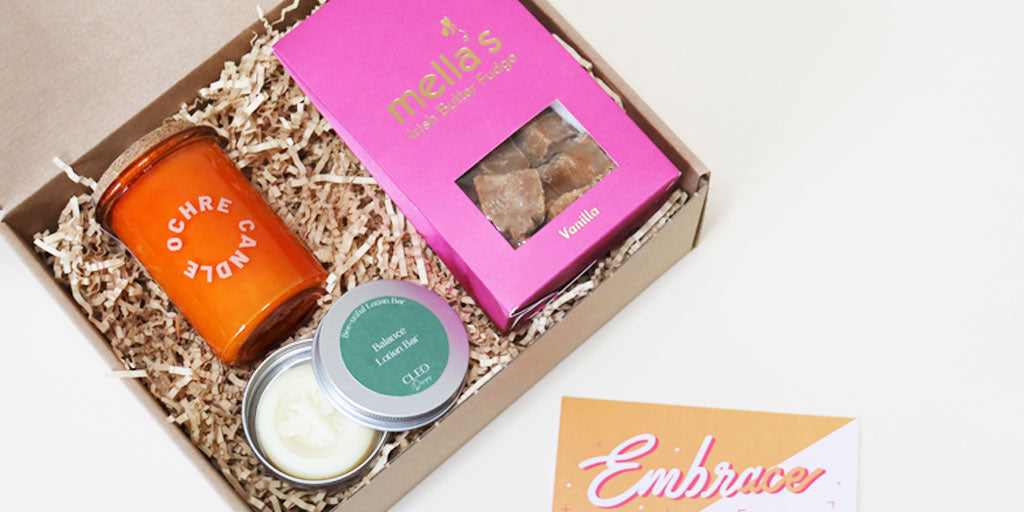 A gift box filled with Irish-made products including a pink fudge box, an orange candle and a bee lotion bar.