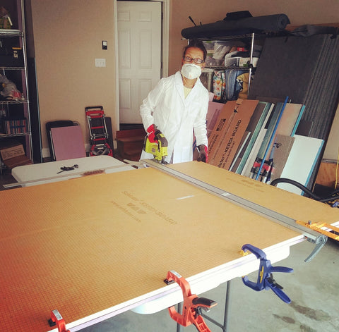 Artist Victoria Primicias wears a mask and lab coat in the garage studio while cutting a board with a jigsaw.